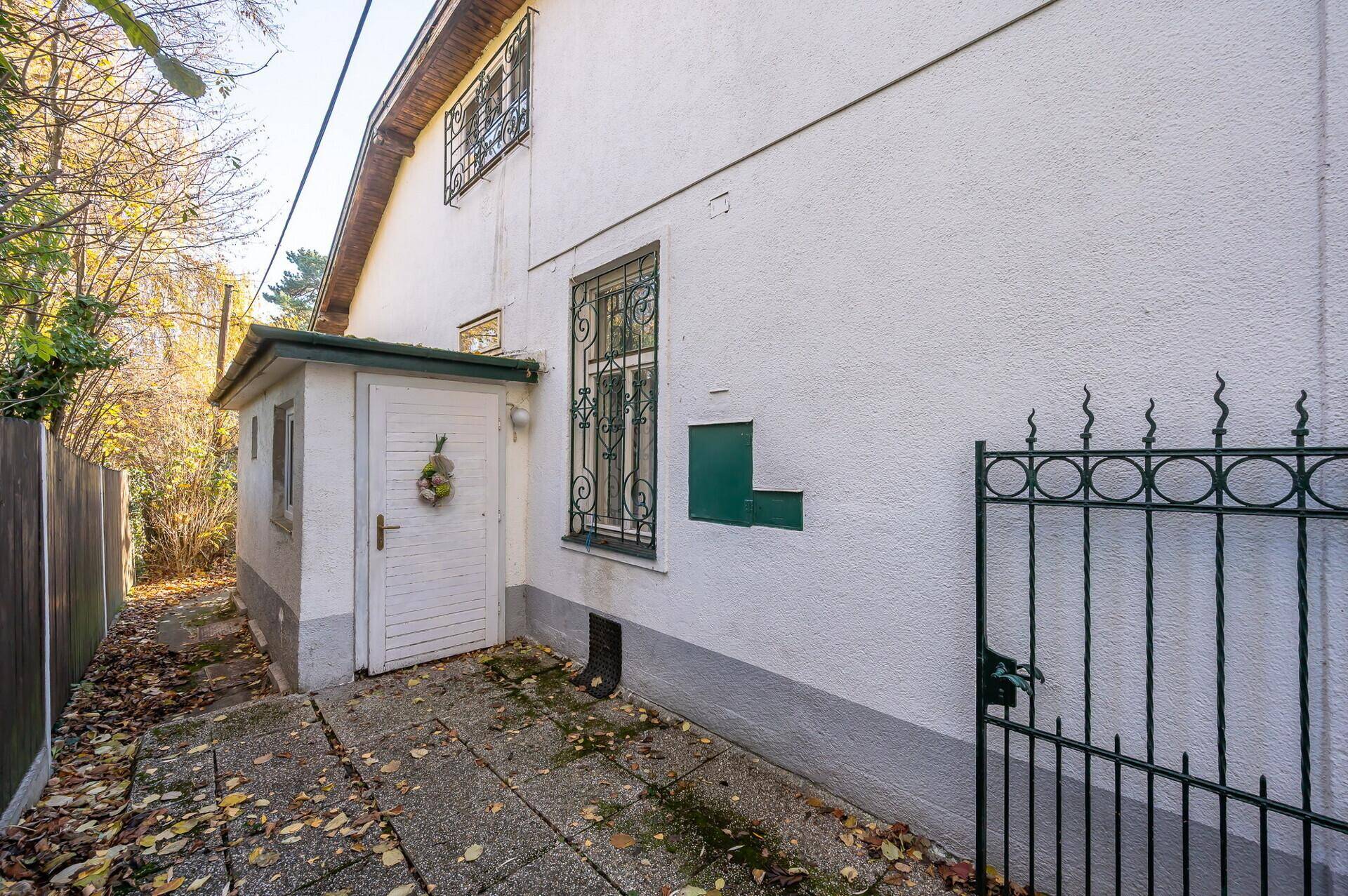 Seitenansicht des Hauses mit Eingangsbereich und schmiedeeisernem Zaun, umgeben von Herbstlaub.