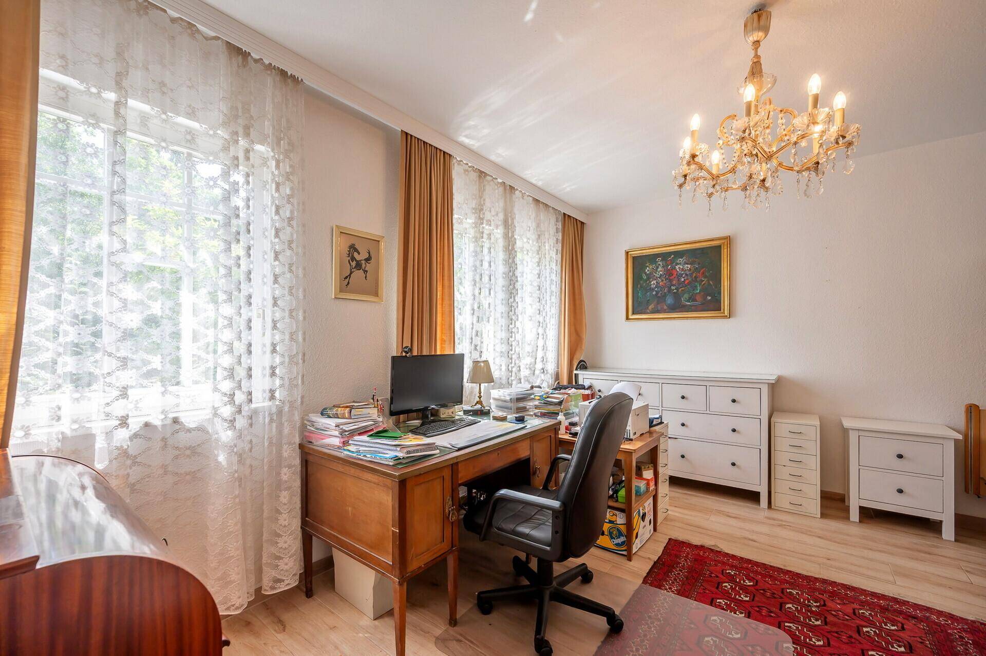 Helles Arbeitszimmer mit Schreibtisch, Kronleuchter und großen Fenstern.