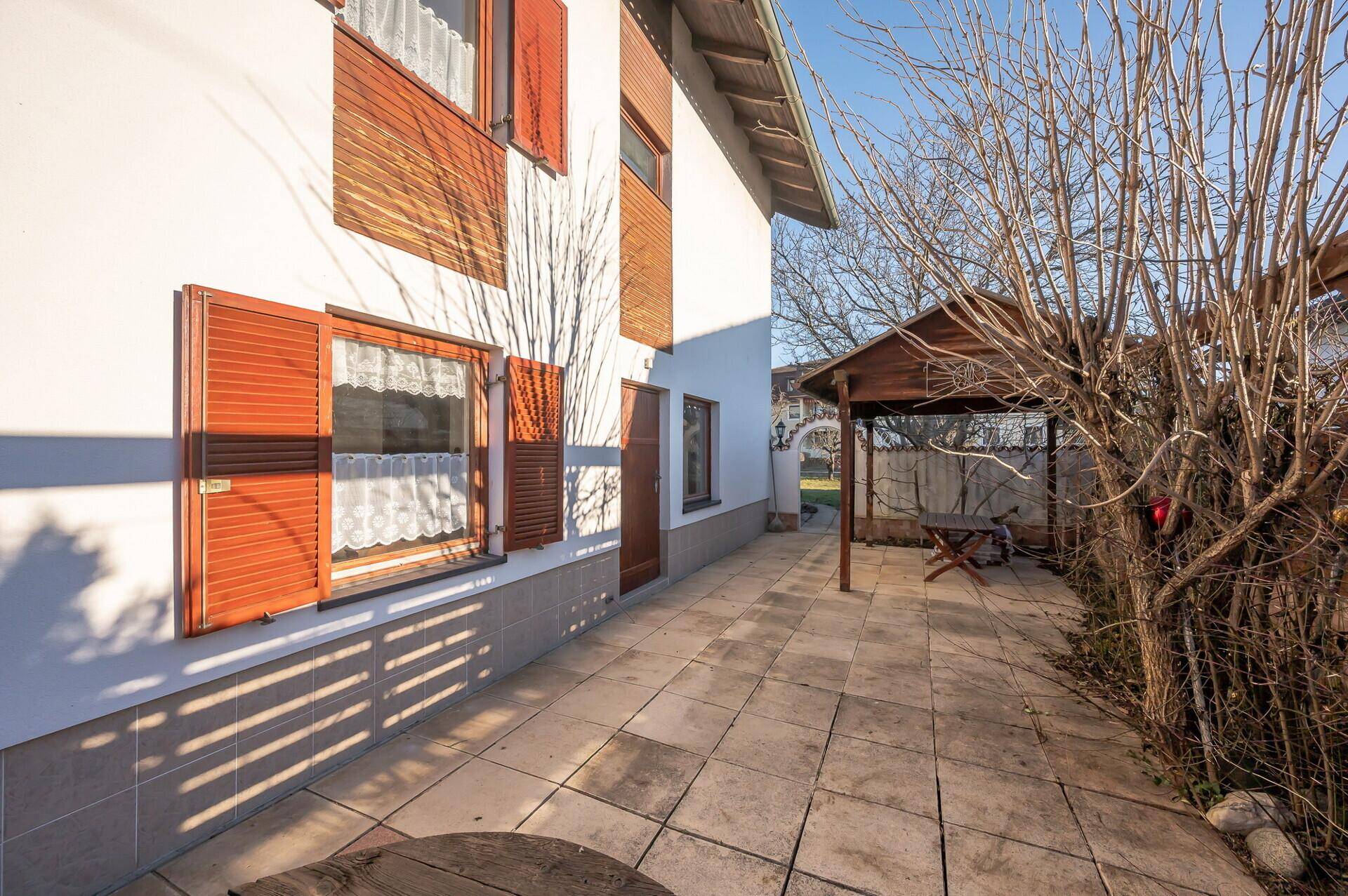 Sonniger Terrassenbereich mit Pflastersteinen, Holzhütte und Pergola neben der Hausfassade.