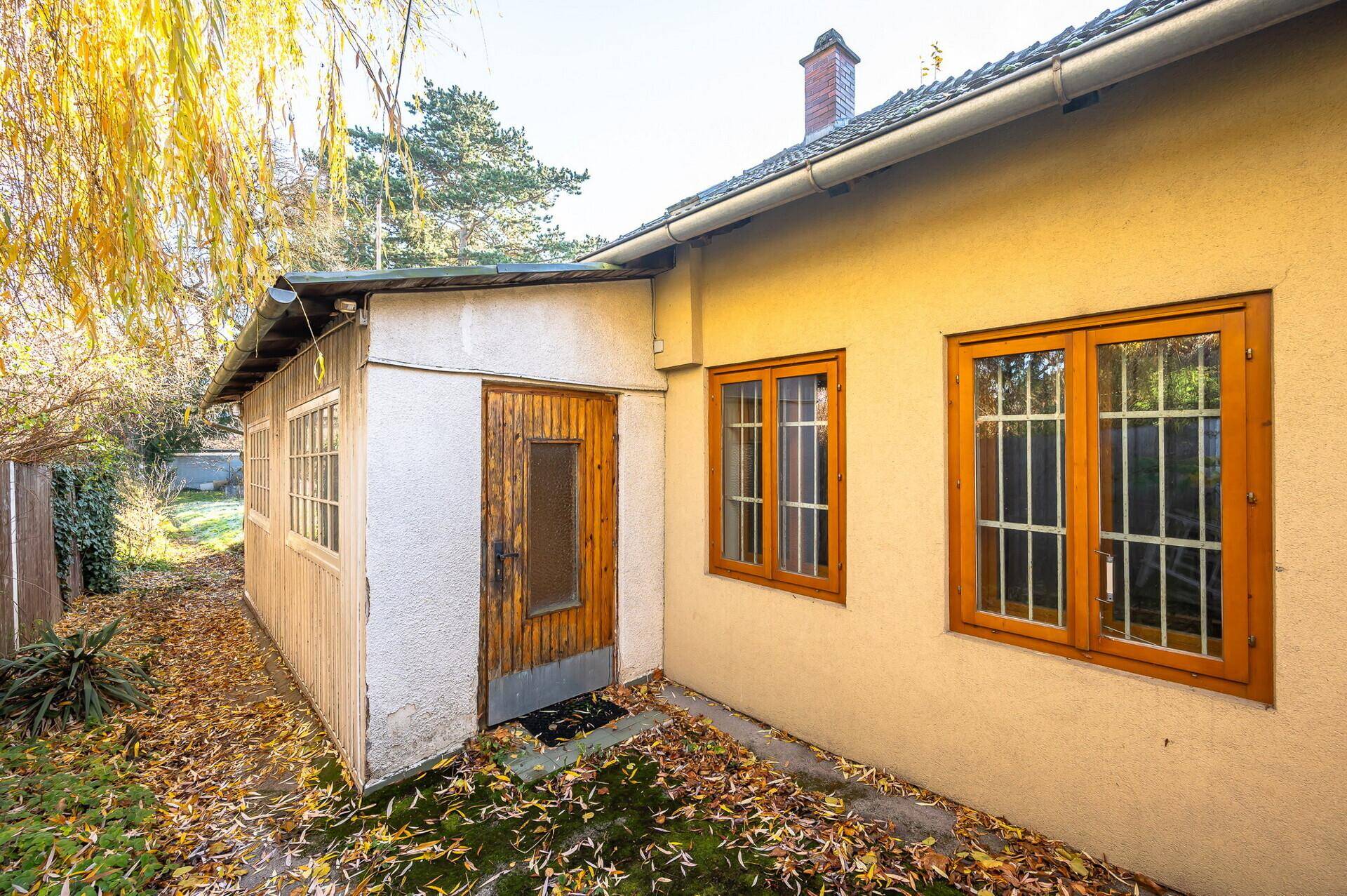 Seitenansicht des Hauses mit Holztüren und Fenstern, umgeben von herbstlichem Laub.