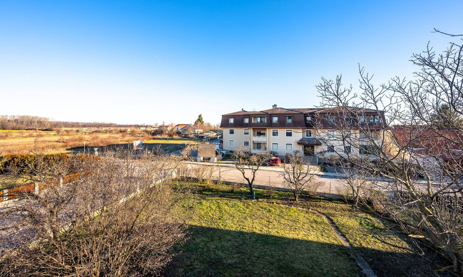 Weitläufiger Blick über grüne Felder und eine Wohnsiedlung unter klarem blauem Himmel.