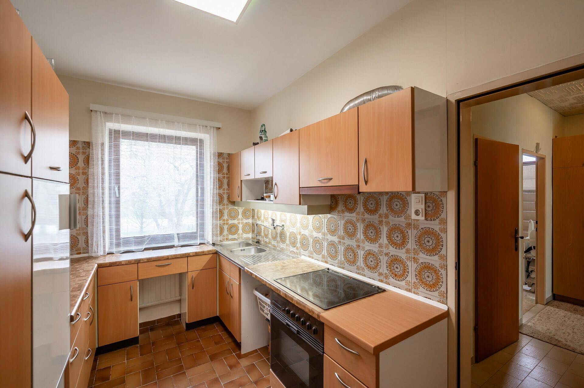 Funktionale Küche mit Holzschränken, Fliesenboden und Fenster mit Blick ins Freie.