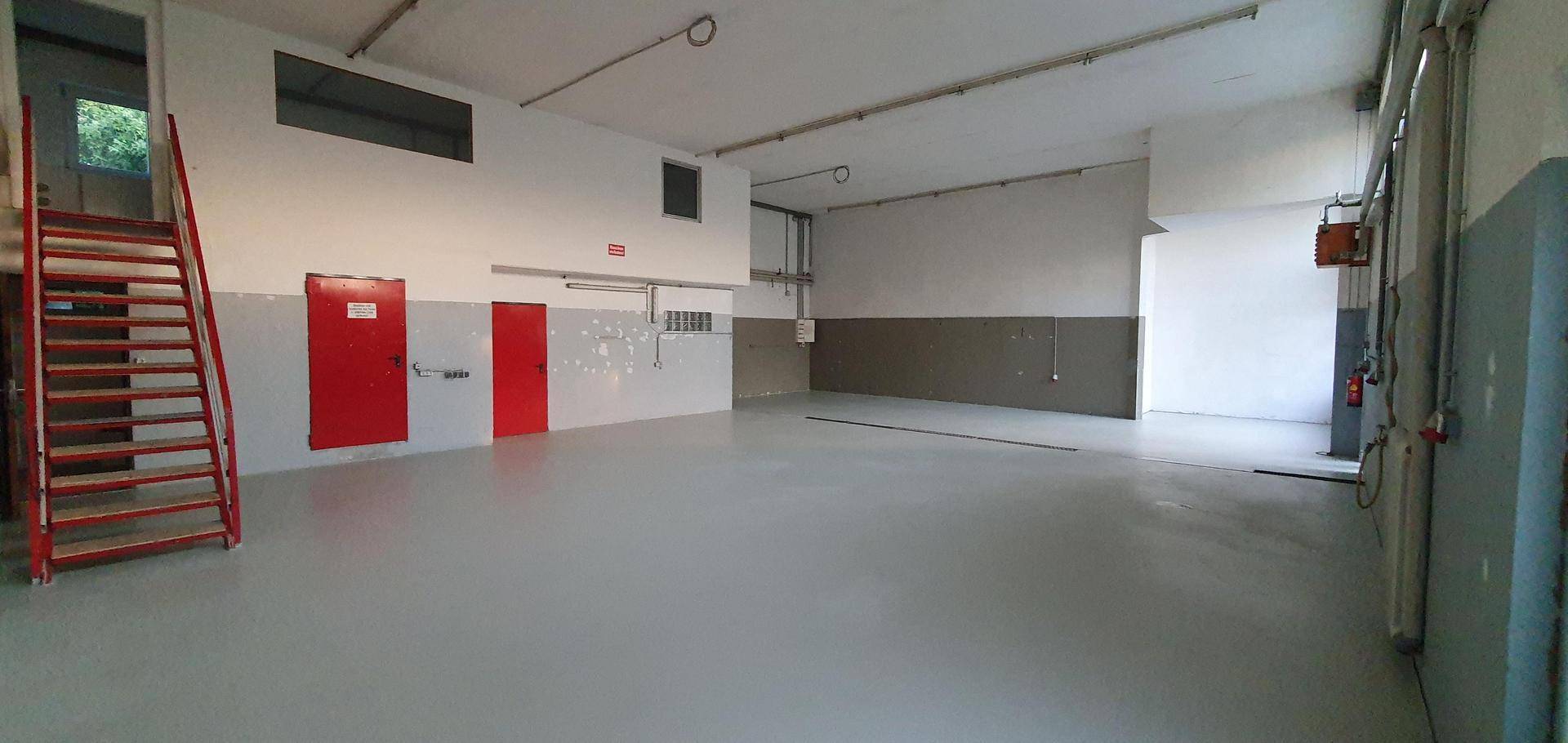 Leere Werkshalle mit einer roten Stahltreppe, zwei roten Türen und einem hellgrauen Boden.