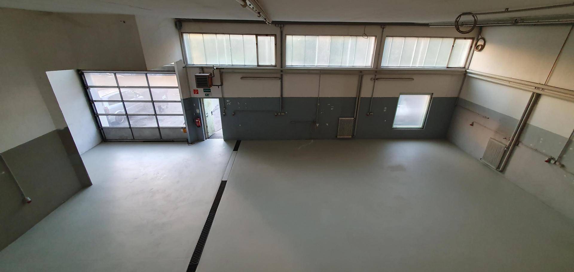 Große, leere Werkshalle mit Rolltor und hohen Fenstern, die viel Tageslicht hereinlassen.