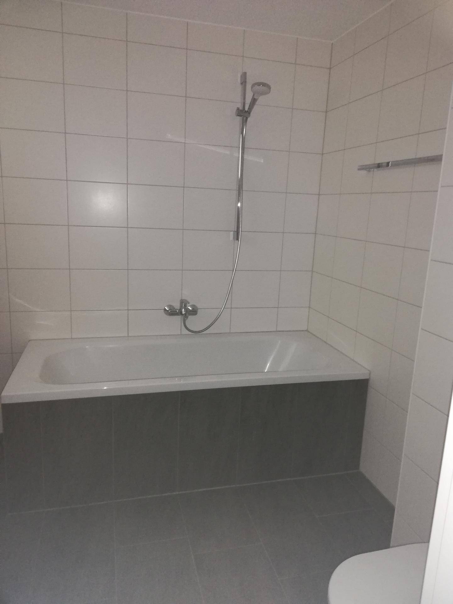 Badezimmer mit weißen Wandfliesen, grauen Bodenfliesen und einer Badewanne mit Duschfunktion.