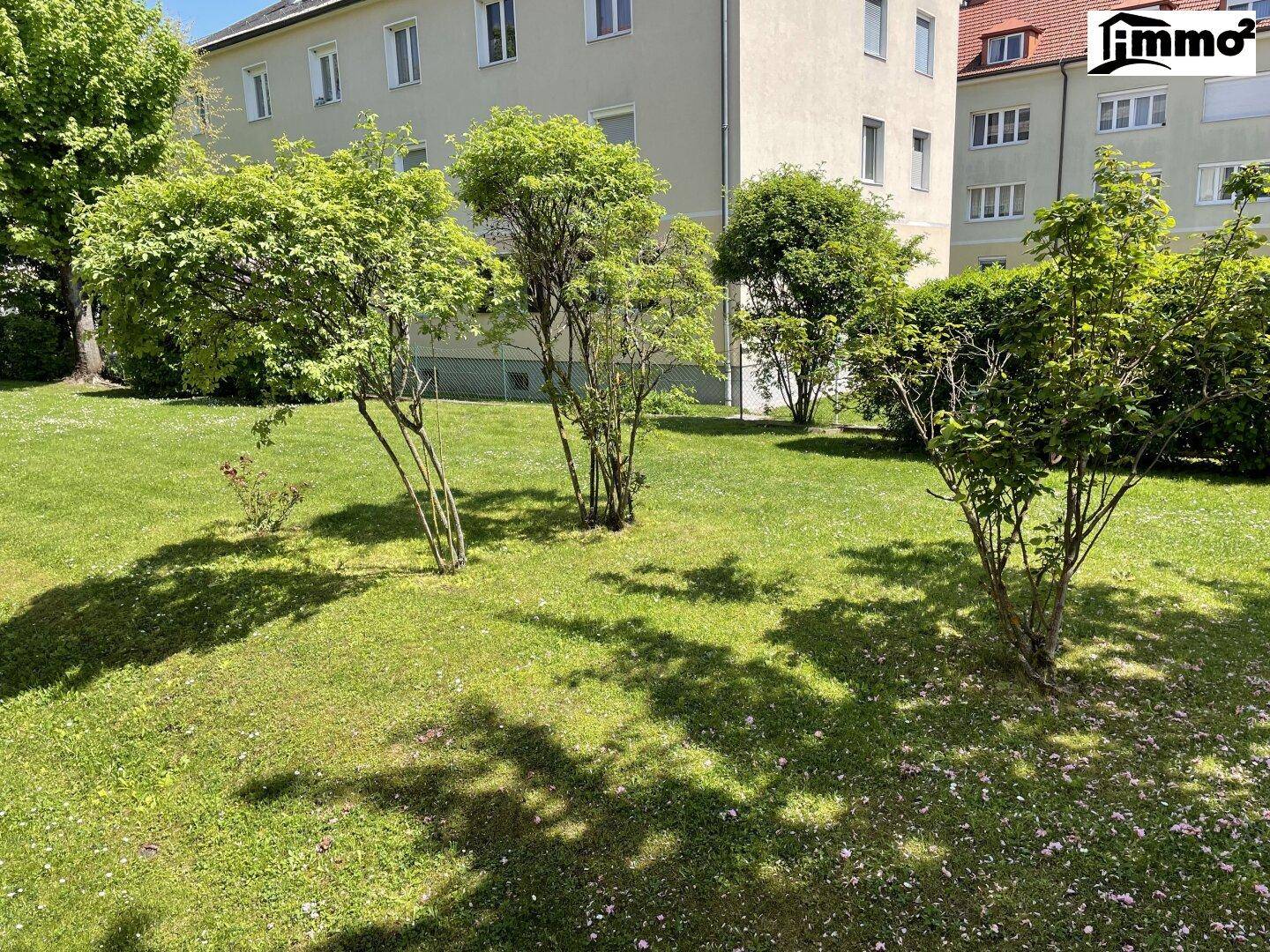 Weitläufige Rasenfläche mit jungen Bäumen und Sträuchern vor einem Wohngebäude an einem hellen Tag.