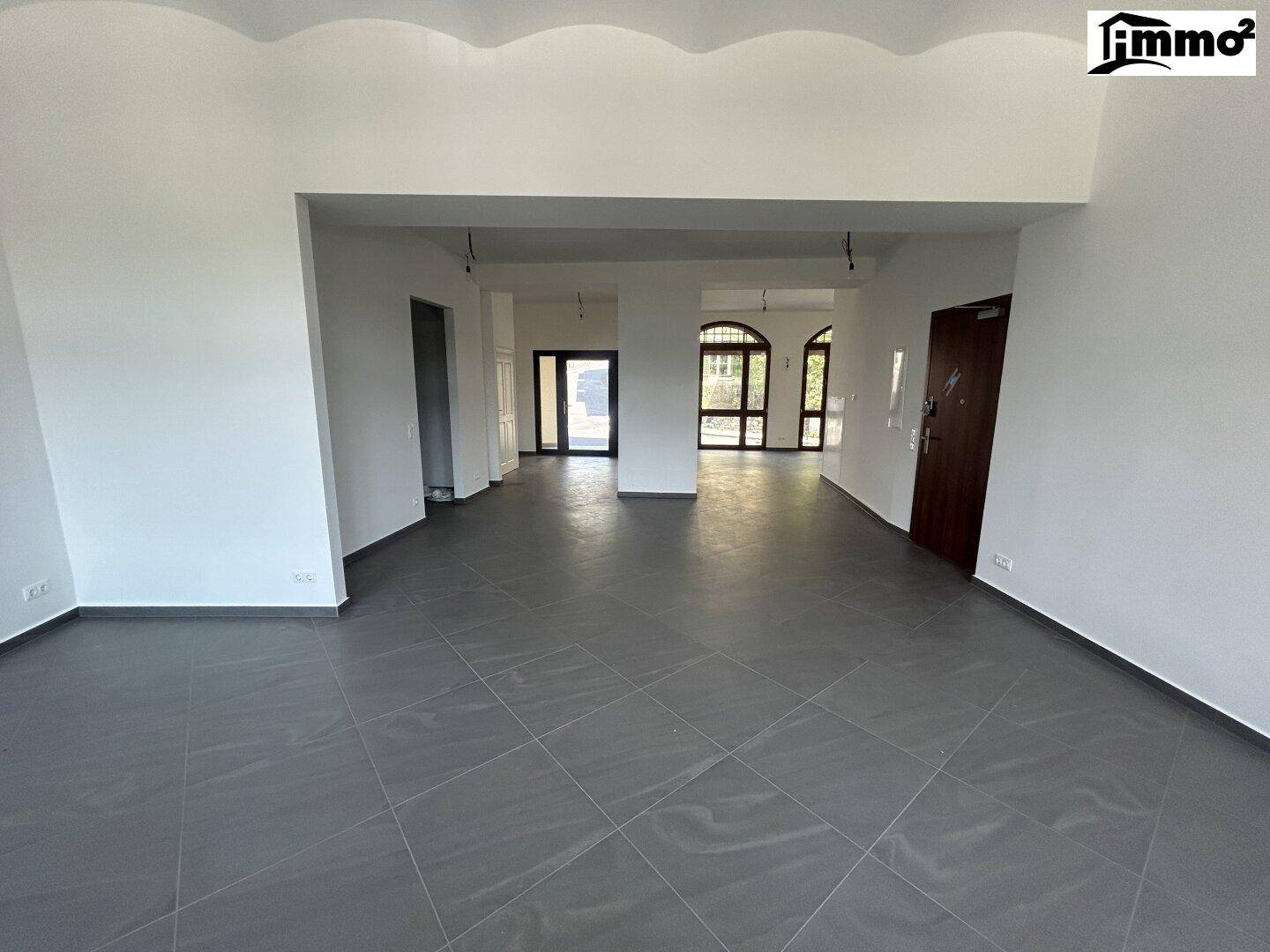 Großer, leerer Innenraum mit dunklem Fliesenboden, weißen Wänden und mehreren Bogenfenstern im Hintergrund.