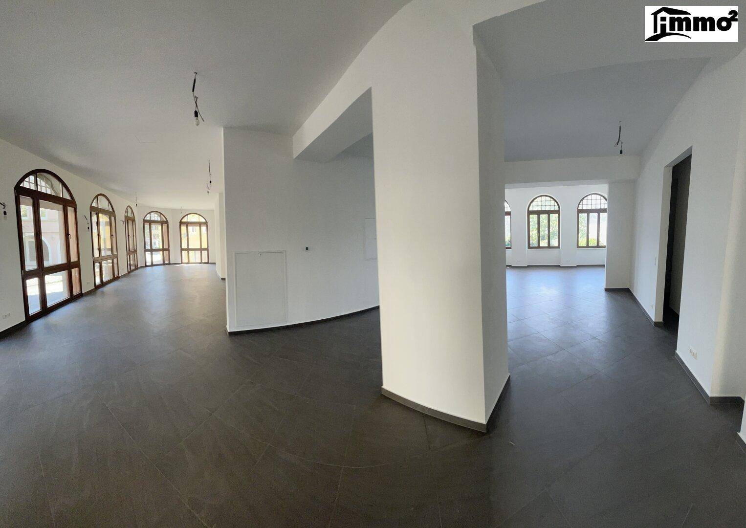 Weitläufiger, leerer Raum mit dunklem Fliesenboden, weißen Wänden und Säulen, Bogenfenster im Hintergrund.