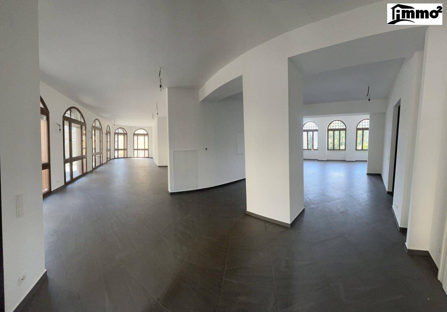 Weitläufiger, leerer Raum mit dunklem Fliesenboden, weißen Wänden und Säulen, Bogenfenster im Hintergrund.