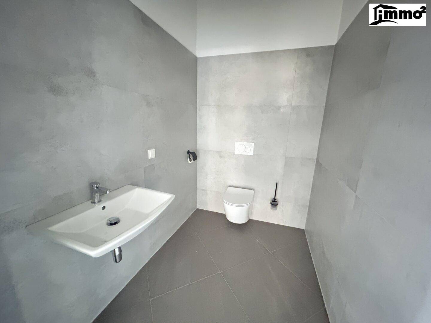 Modernes Gäste-WC mit grauen Fliesen an Boden und Wänden, wandhängendem WC und weißem Waschbecken.