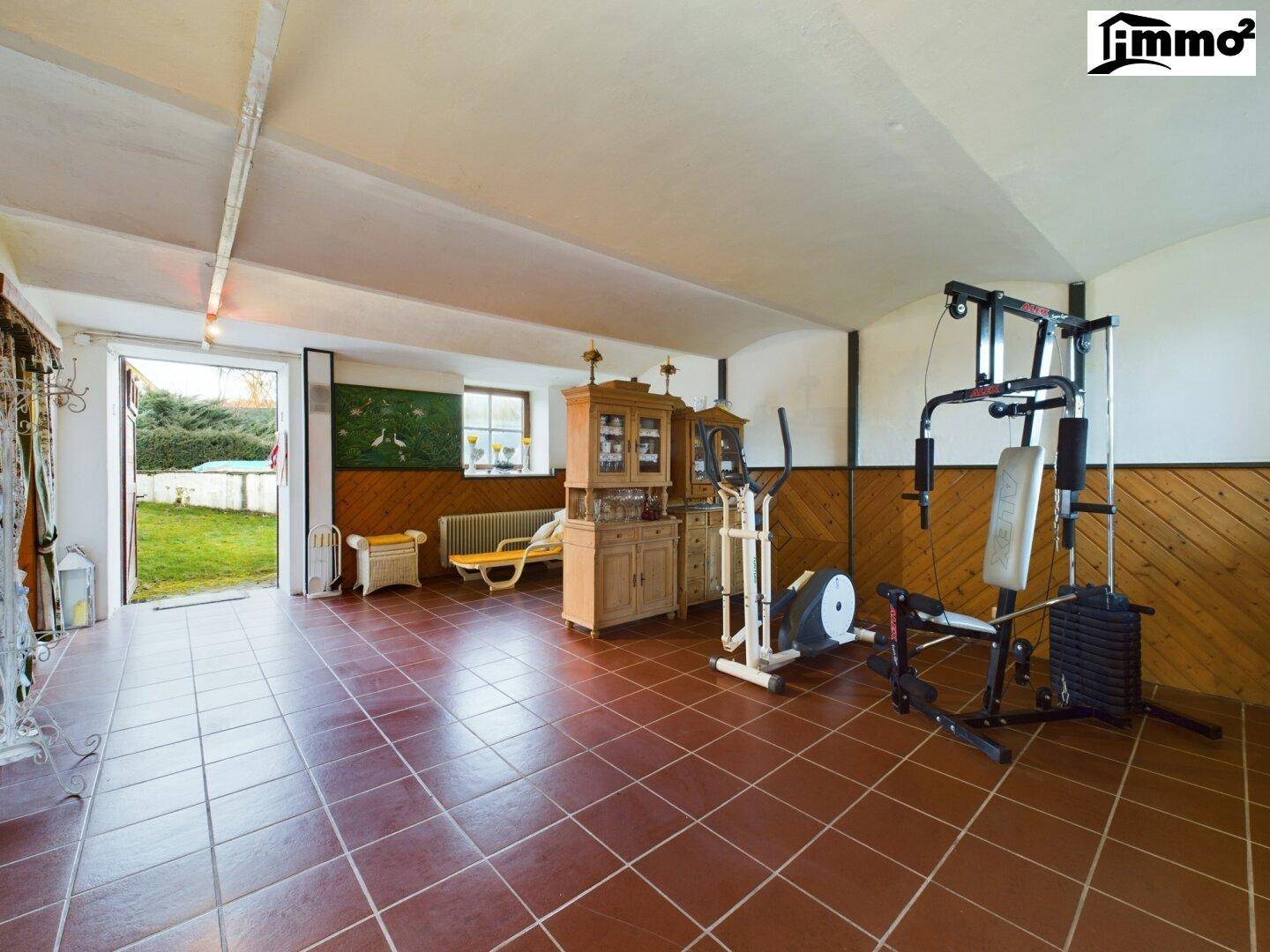 Großer Fitnessraum mit Sportgeräten, Fliesenboden und Zugang zum Gartenbereich.