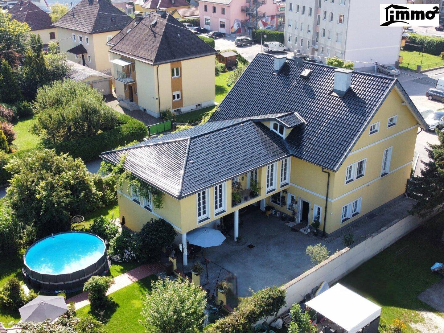 Luftaufnahme eines gelben Hauses mit schwarzem Dach, Garten und Pool in einer Wohngegend.