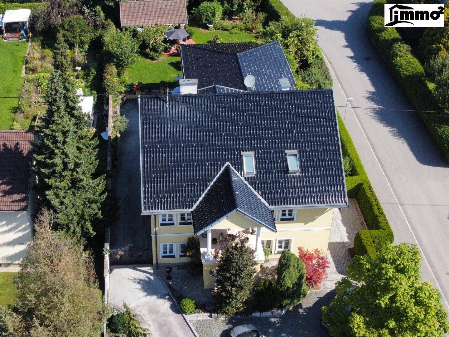 Vogelperspektive des gelben Hauses mit dunklem Dach, umgeben von Bäumen und einer Straße.