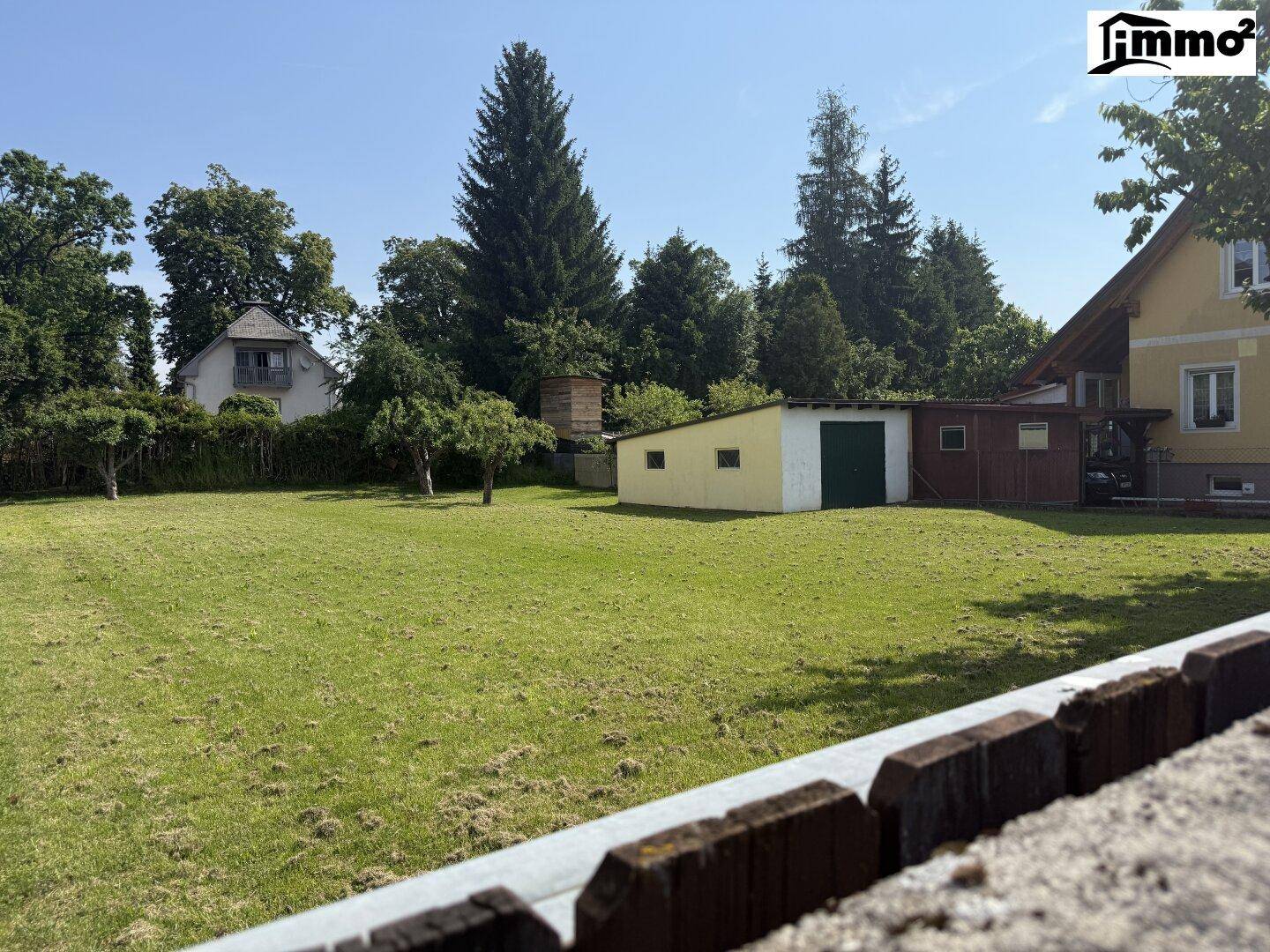Heller Gartenbereich mit Rasen, einem Nebengebäude und umgebenden Bäumen, gesehen über eine niedrige Mauer.