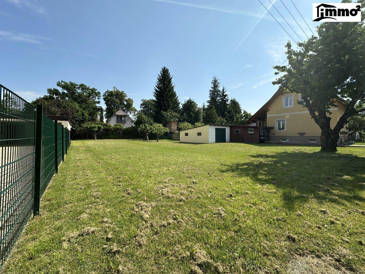 Weitläufiger Gartenbereich mit gepflegtem Rasen, Zaun und einem kleinen Nebengebäude unter blauem Himmel.