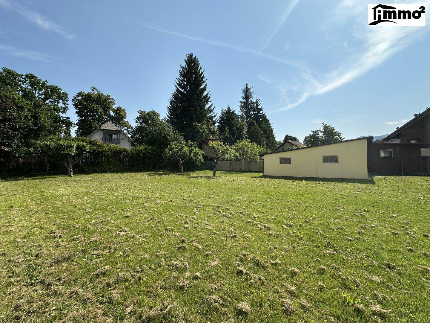 Sonniger Garten mit weitläufiger Rasenfläche, hohen Bäumen und einem kleinen Nebengebäude im Hintergrund.