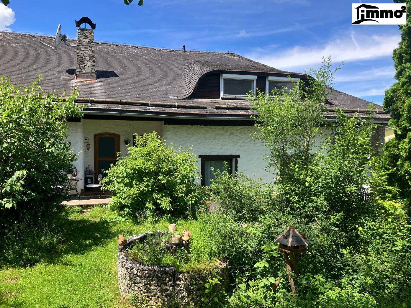 Charmantes Haus mit Mansarddach und naturnahem Gartenbereich unter blauem Himmel.