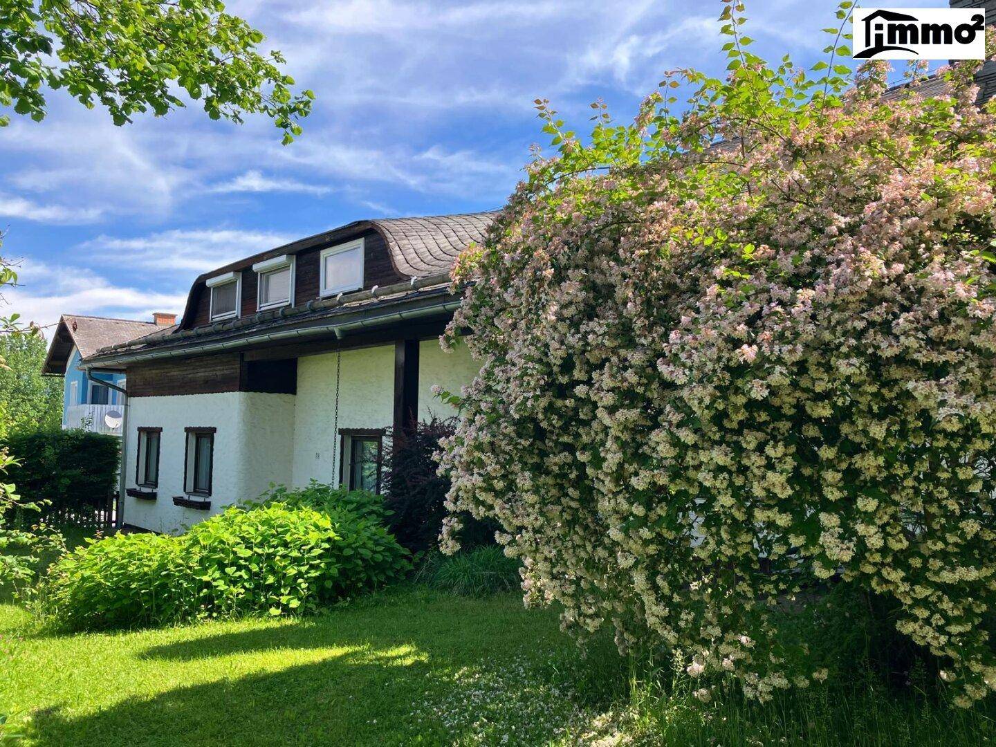 Hausfassade mit blühendem Strauch im Vordergrund und gepflegtem Garten unter sonnigem Himmel.