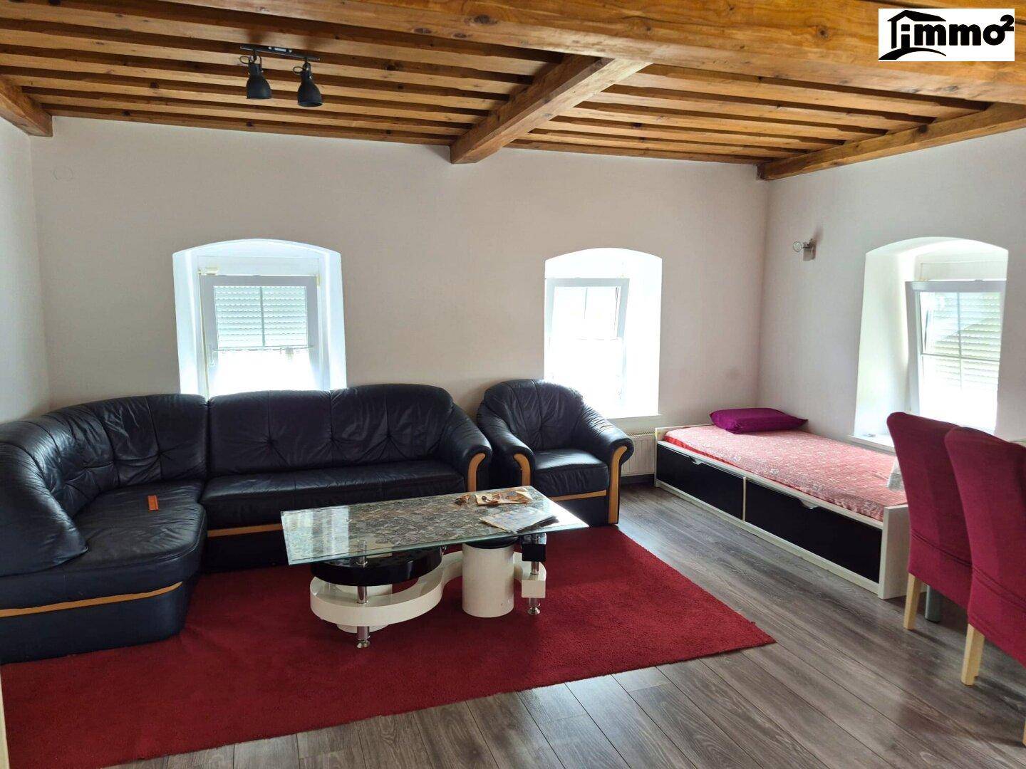 Geräumiges Wohnzimmer mit sichtbaren Holzbalken, dunklem Sofa und rotem Teppich.