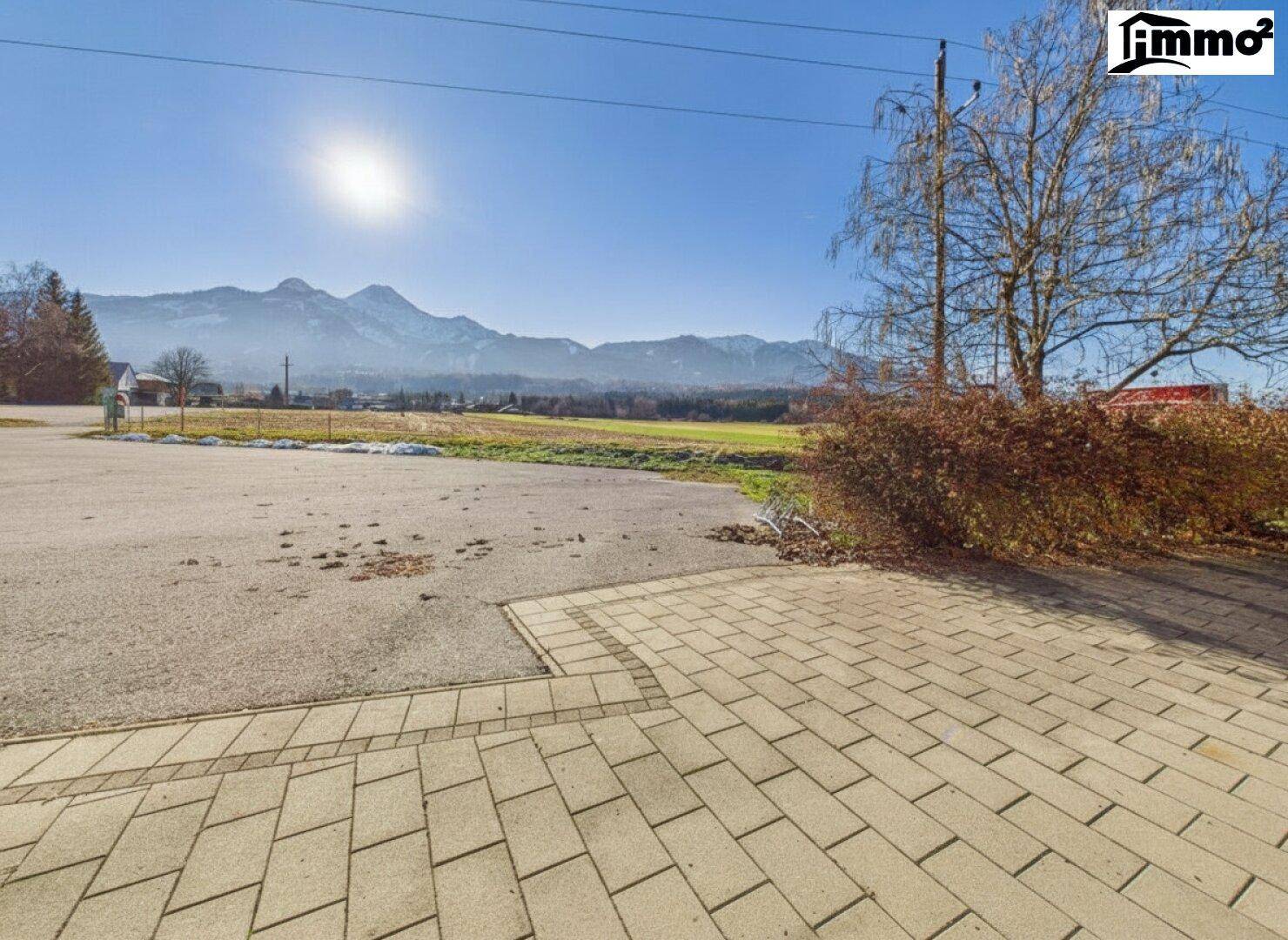 Gepflasterter Bereich neben dem Gebäude mit weitem Blick über grüne Felder und die umliegenden Berge.