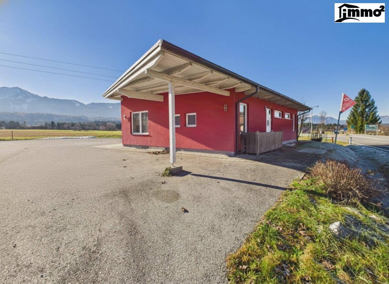 Rotes Gebäude mit überdachtem Eingangsbereich und großem Parkplatz vor einer Bergkulisse.