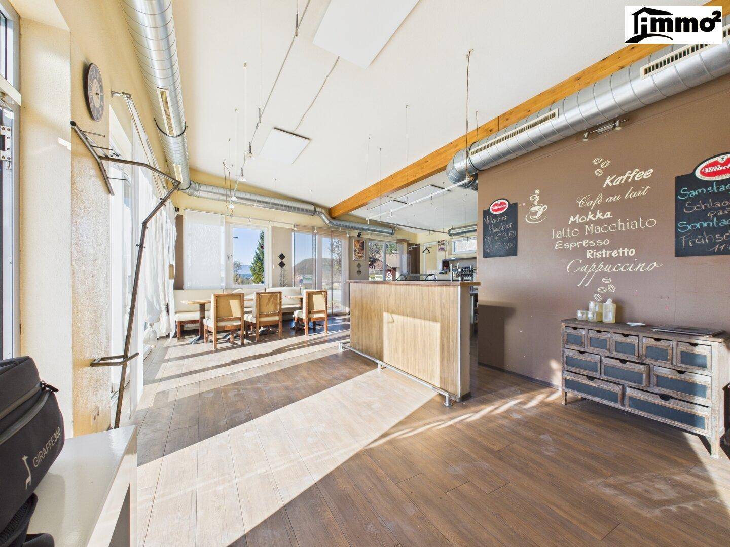 Heller Innenraum mit Holzboden, sichtbaren Lüftungsrohren und großen Fenstern, ideal für ein Café.