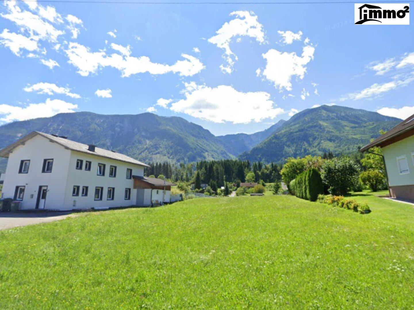 Weitläufige grüne Landschaft mit traditionellen Häusern und beeindruckendem Bergpanorama im Hintergrund.