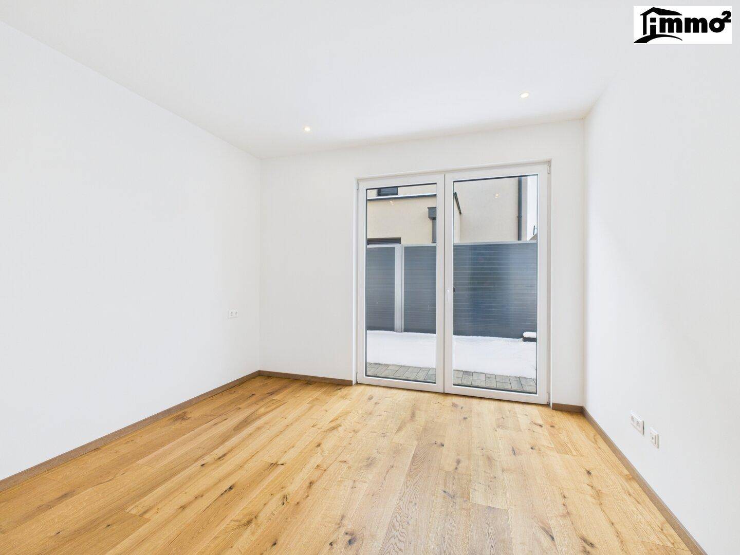 Leeres Zimmer mit Holzboden und großer Fenstertür, die viel Tageslicht hereinlässt.