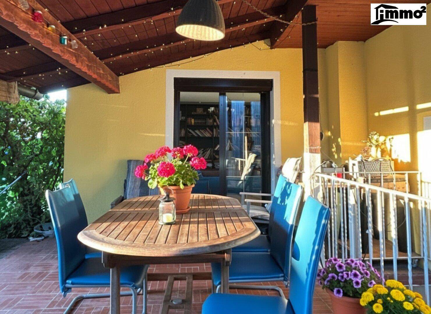 Gemütliche überdachte Terrasse mit Holztisch und blauen Stühlen.