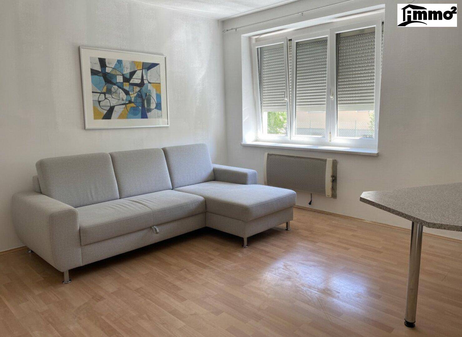 Gemütliches Wohnzimmer mit grauem Ecksofa, Laminatboden und Fenster mit Rollläden.