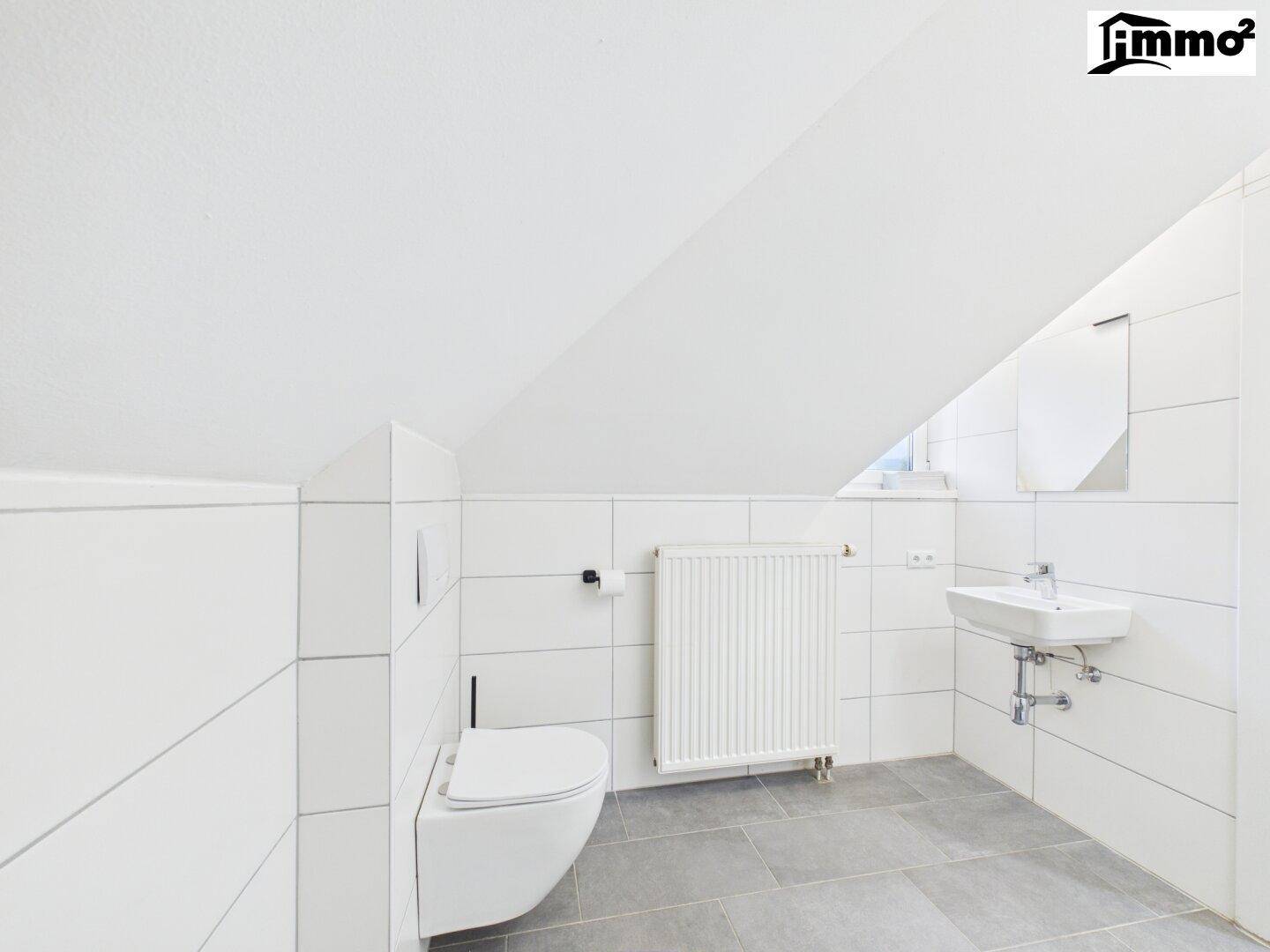 Modernes WC mit Wand-WC, Waschbecken und Fliesenboden unter Dachschräge, inklusive Fenster für Tageslicht.