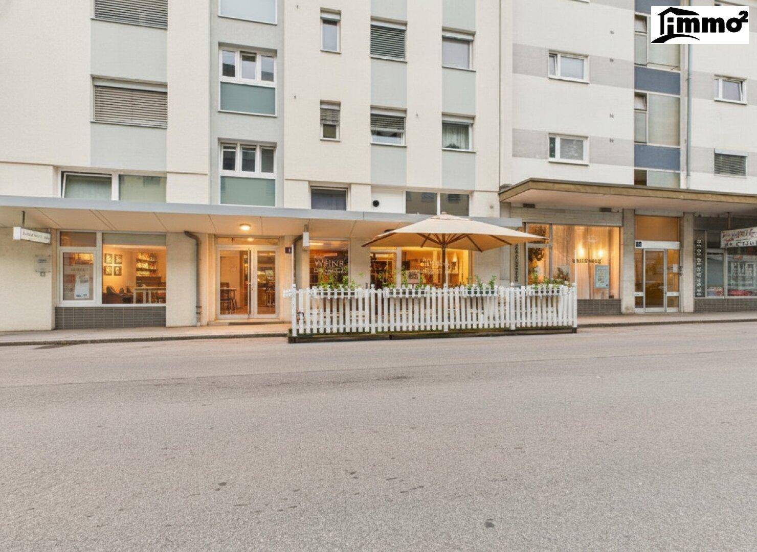 Gebäudeansicht von der Straße mit Außenterrasse und umliegenden Geschäften.