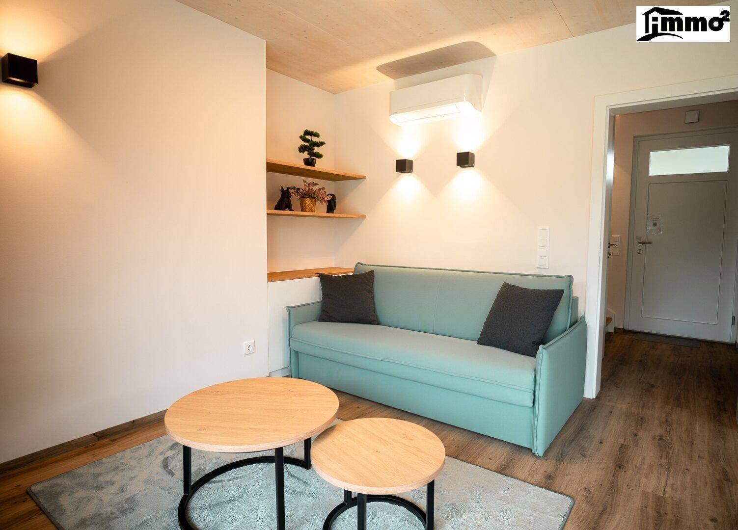 Heller Wohnbereich mit mintgrünem Sofa, Holztischen und Wandregalen, gut beleuchtet.