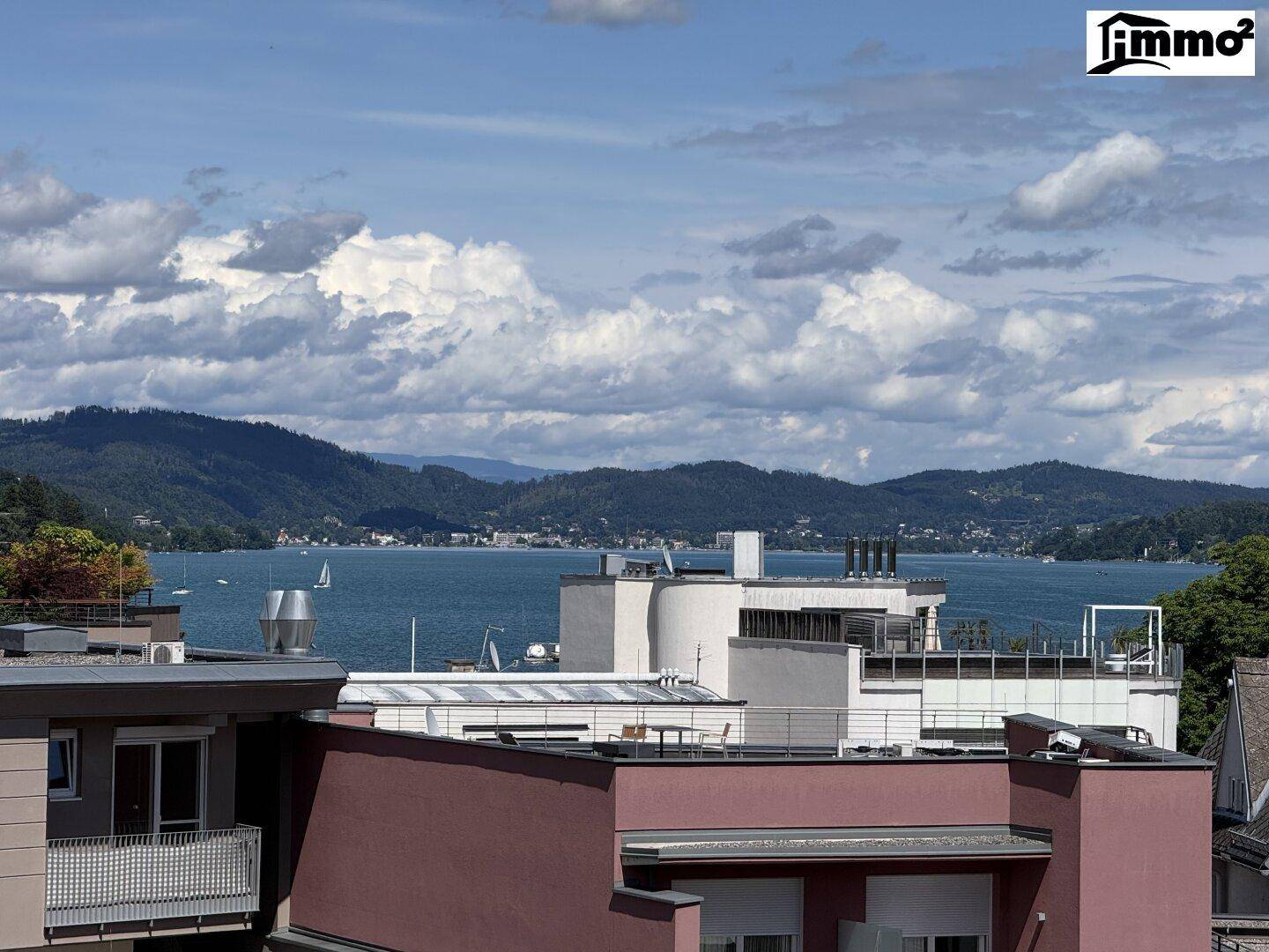 Weitläufiger Blick auf den See mit Segelbooten und bewaldeten Bergen unter einem bewölkten Himmel.
