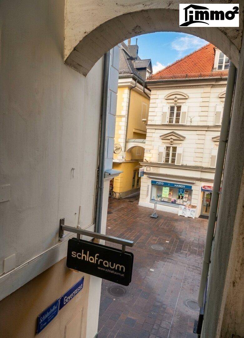 Blick durch einen Torbogen auf eine historische Gasse mit Geschäften und Gebäuden im Stadtzentrum.