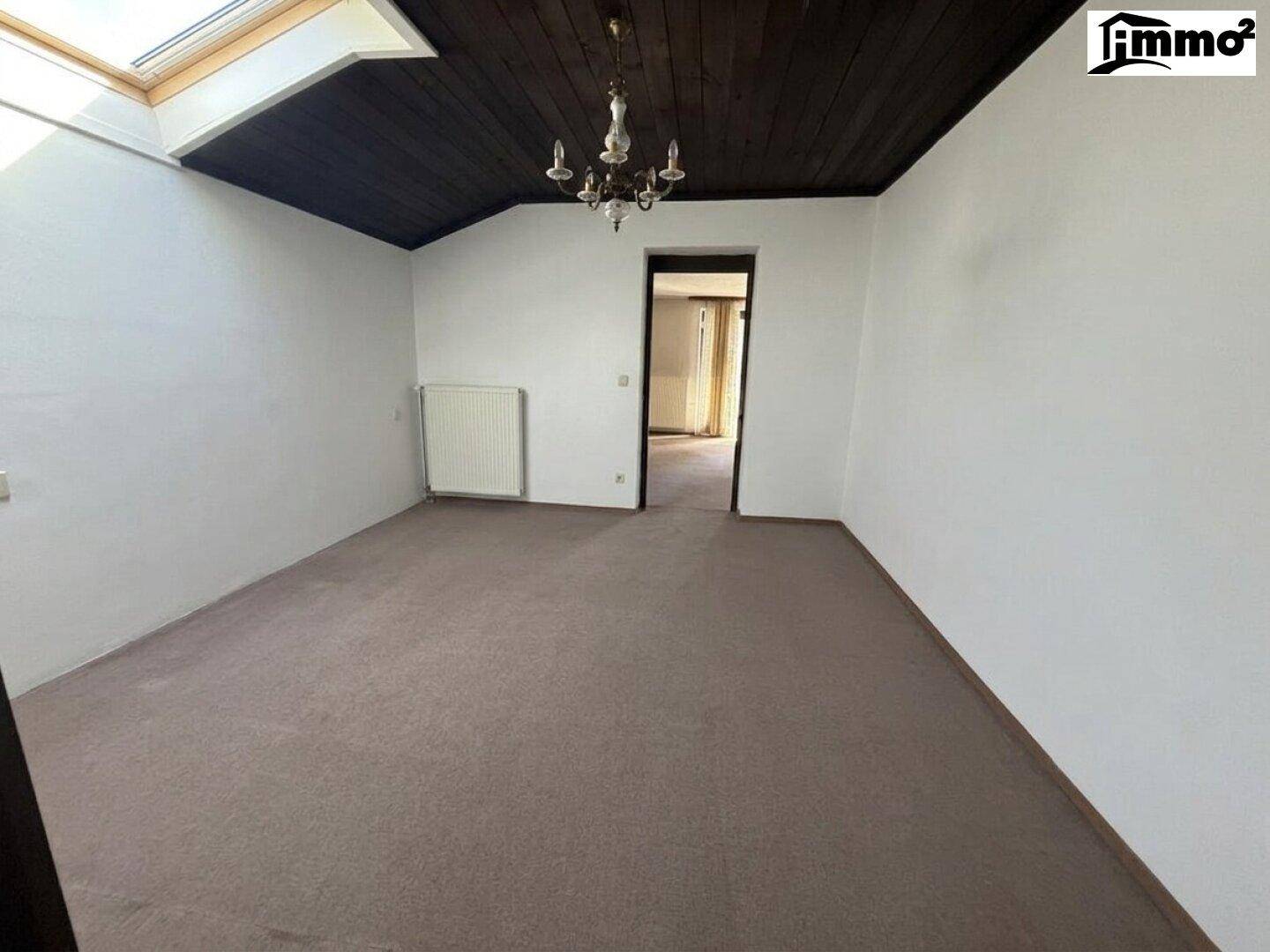 Leerer Raum mit Teppichboden, dunkler Holzdecke, Kronleuchter und Dachfenster.