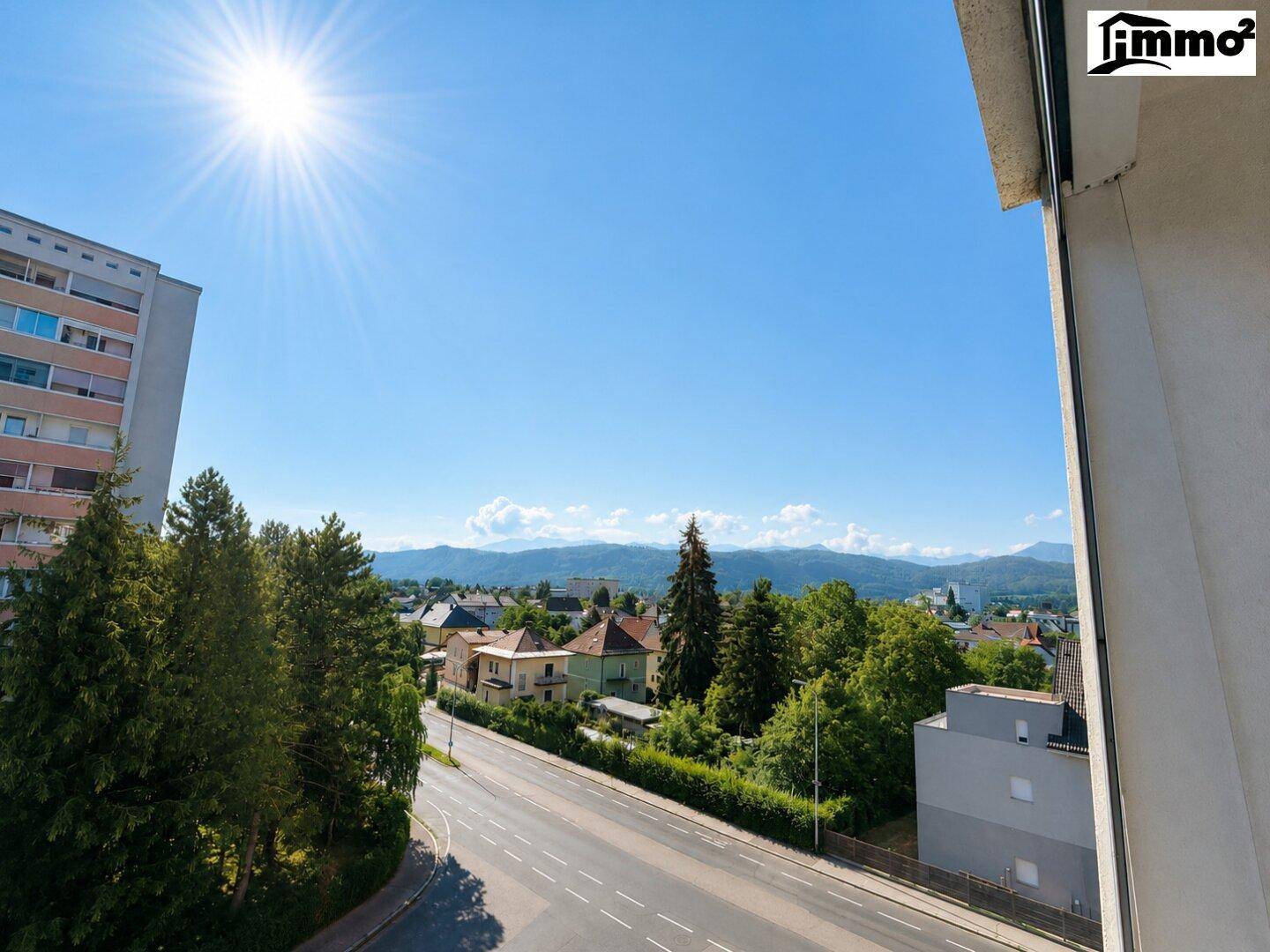 Weitläufiger Blick über die Stadt, grüne Bäume und ferne Berge unter strahlendem Himmel.