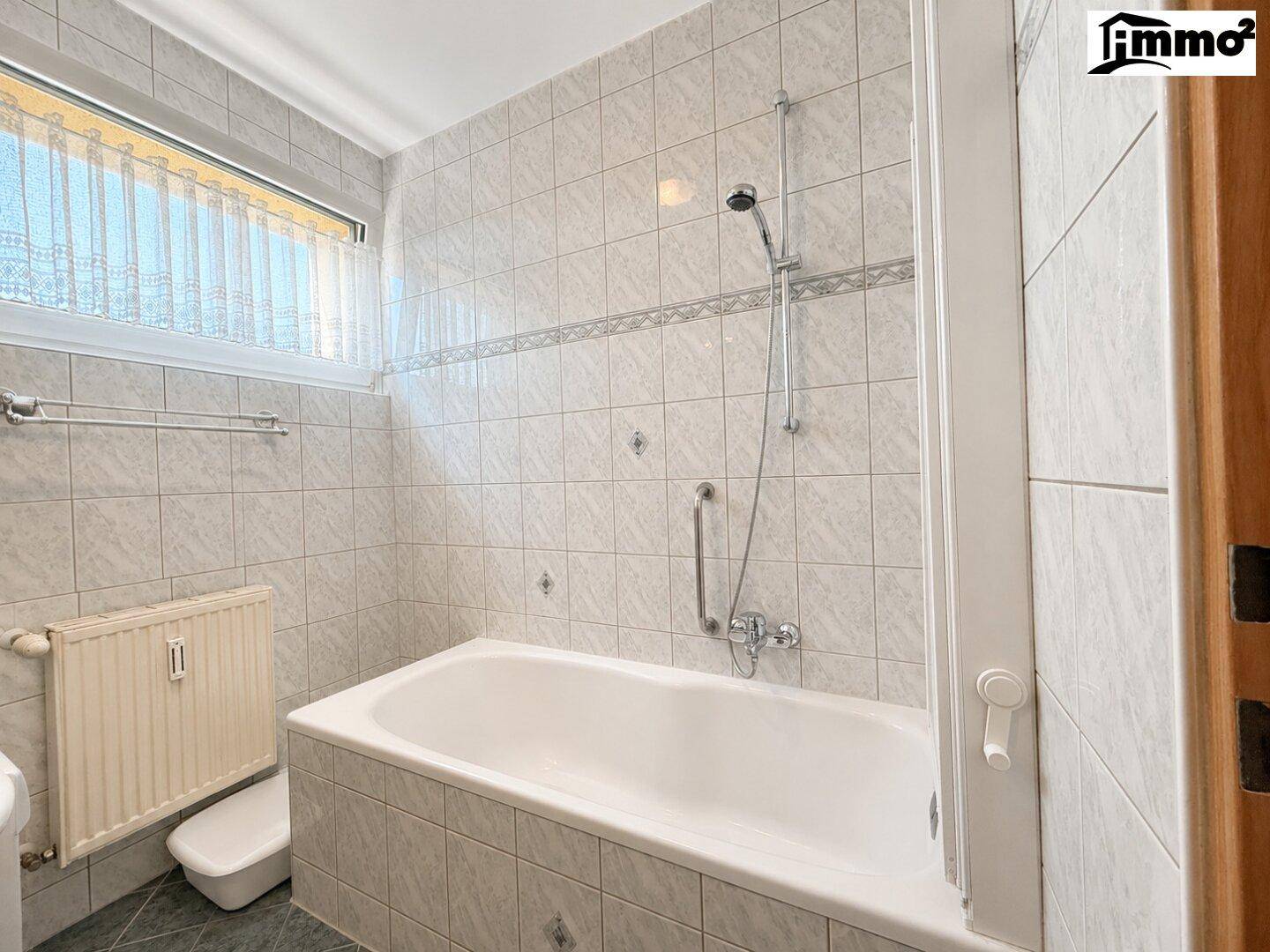 Gefliestes Badezimmer mit Badewanne, Duschvorrichtung und Fenster für Tageslicht.