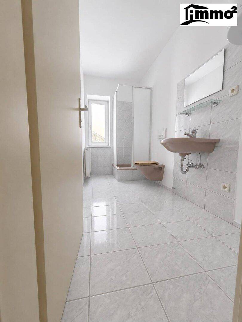 Badezimmer mit hellgrauen Fliesen, Einzelwaschbecken, Dusche und Fenster.