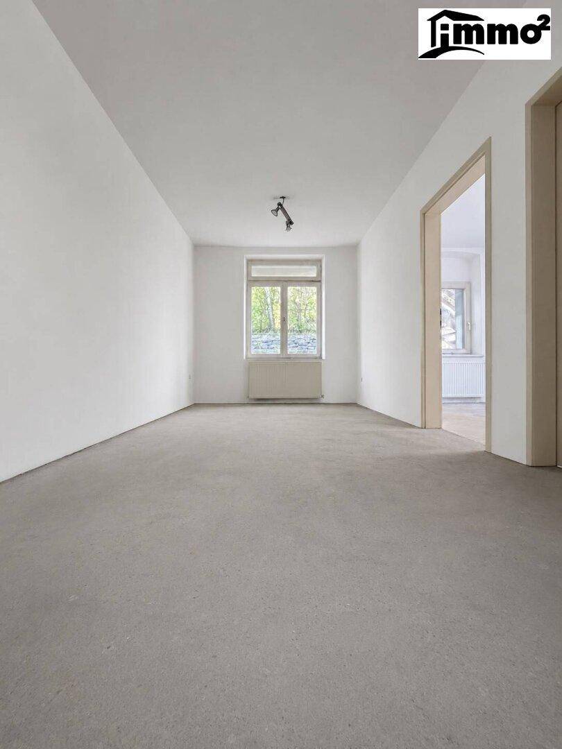 Leerer, heller Raum mit weißen Wänden, Fenster und grauem Bodenbelag.