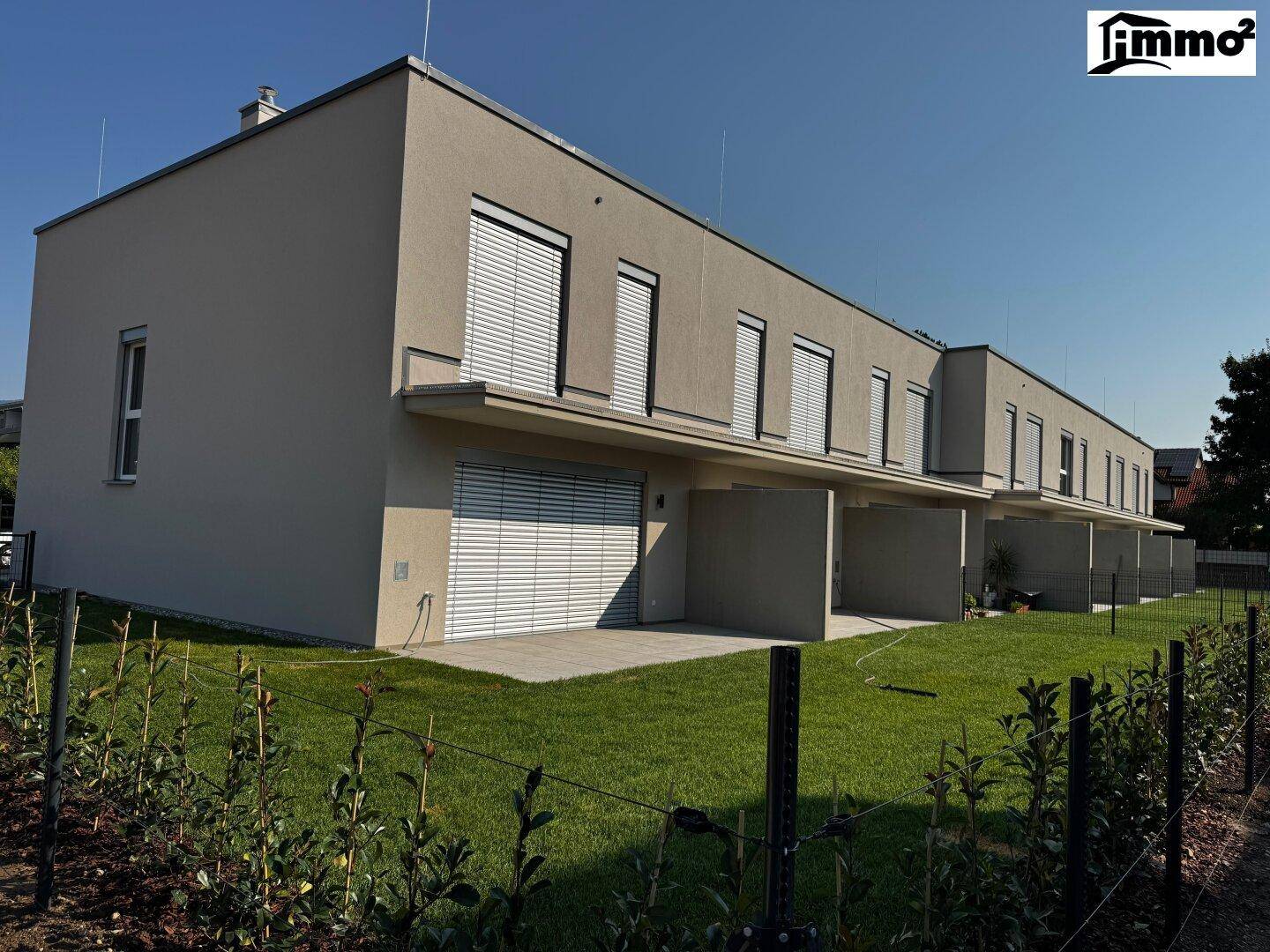 Moderne Reihenhäuser mit heller Fassade, Rollläden und gepflegtem Rasenbereich unter blauem Himmel.