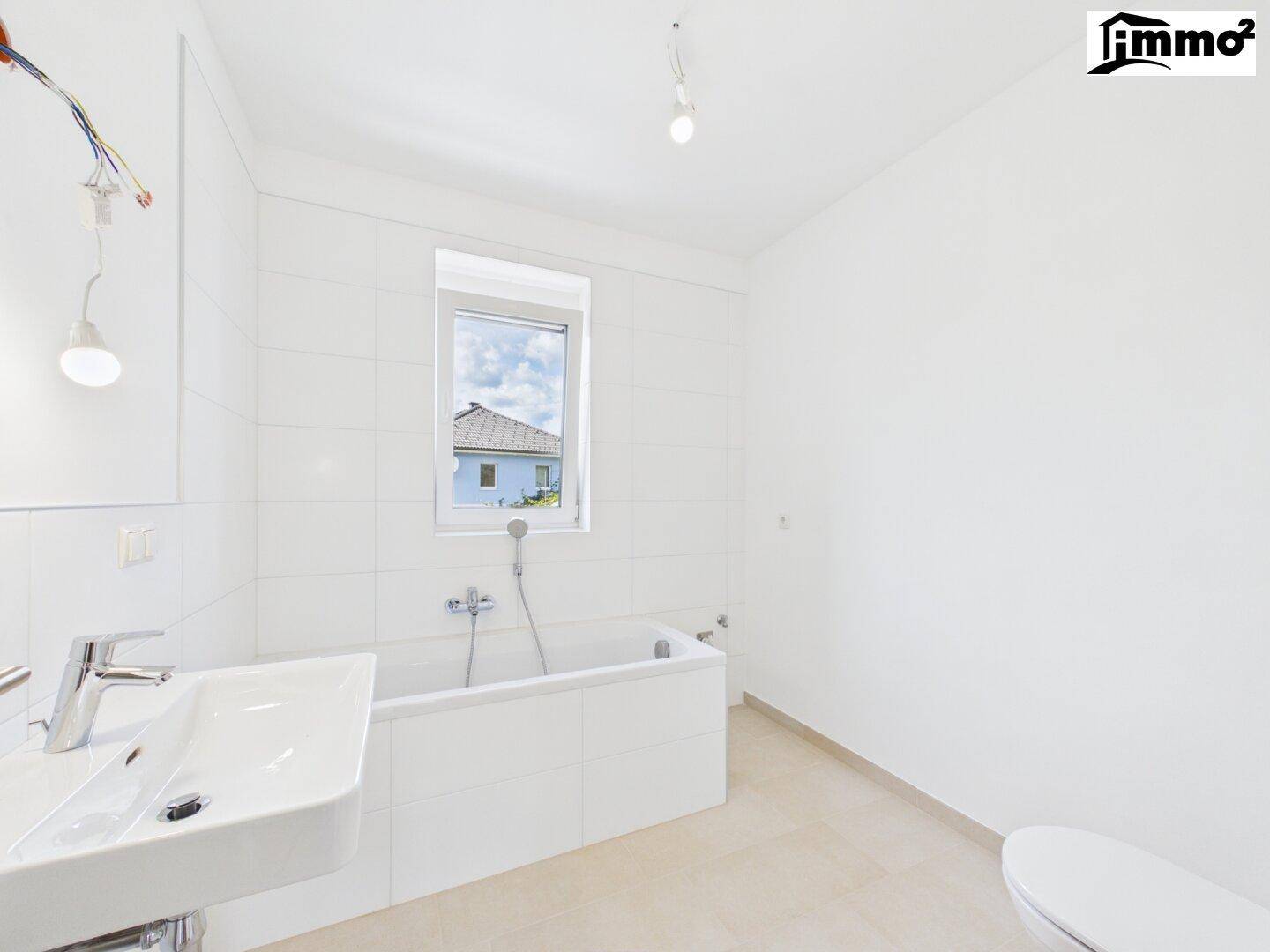 Modernes Badezimmer mit Badewanne, Waschbecken und Fenster, hell gefliest in Weiß und Beige.