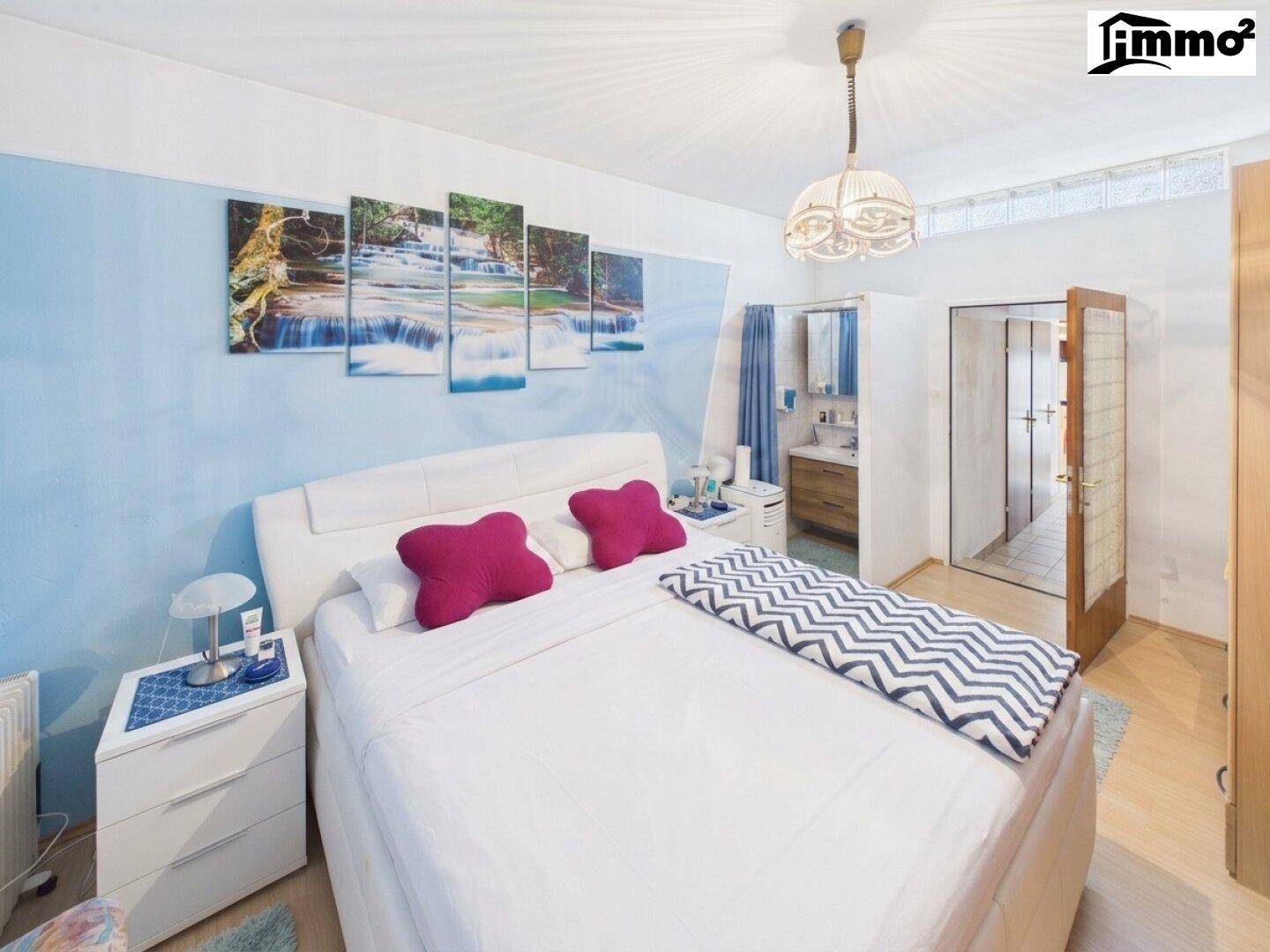 Schlafzimmer mit Doppelbett, blauer Akzentwand und direktem Zugang zu einem kleinen Badbereich.