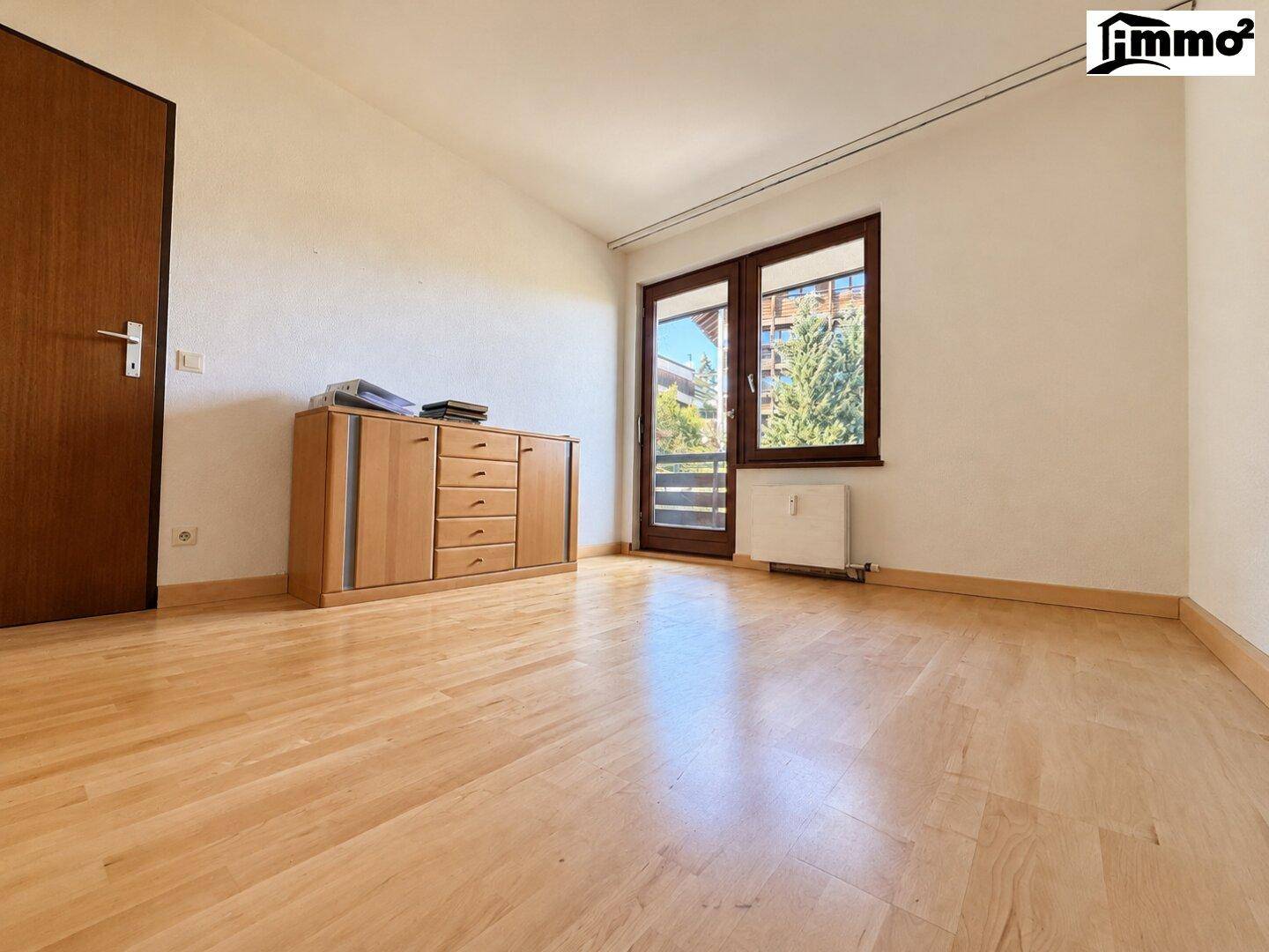 Helles Schlafzimmer mit Holzboden, Kommode und Zugang zu einem Balkon mit Blick ins Grüne.
