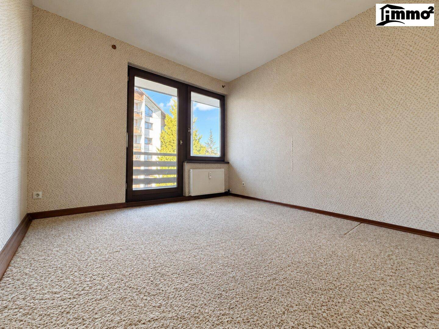 Leeres Zimmer mit Teppichboden, strukturierten Tapeten und Zugang zu einem Balkon.