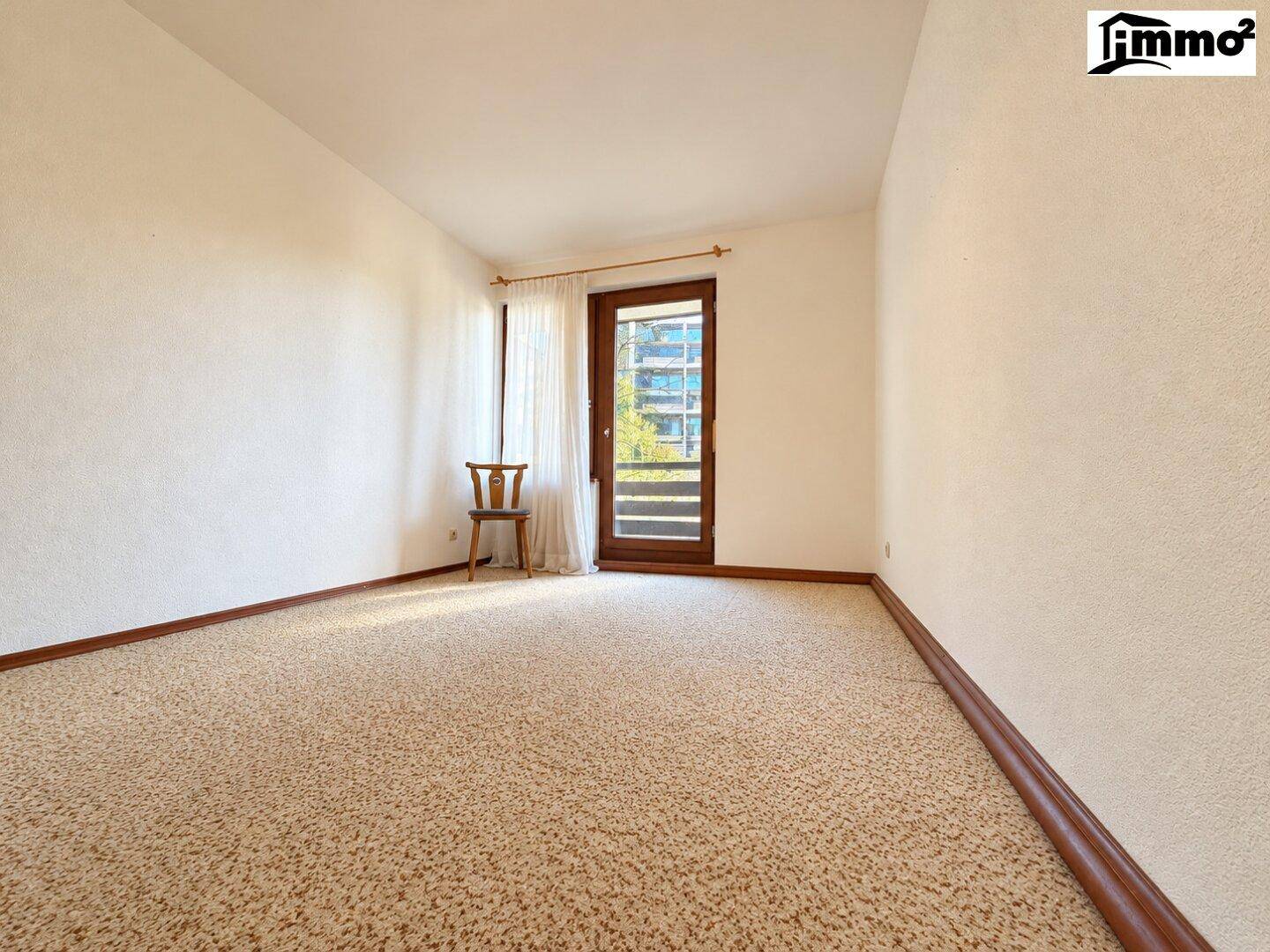 Leeres Schlafzimmer mit Teppichboden, weißen Wänden und Zugang zu einem Balkon.