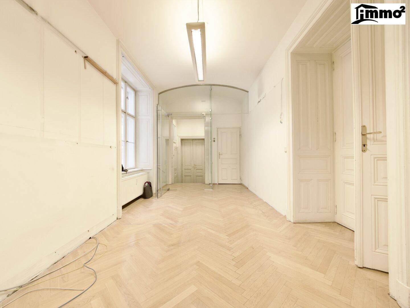 Langer Flur mit Fischgrätparkett, hohen Decken und einem großen Fenster, das viel Tageslicht hereinlässt.