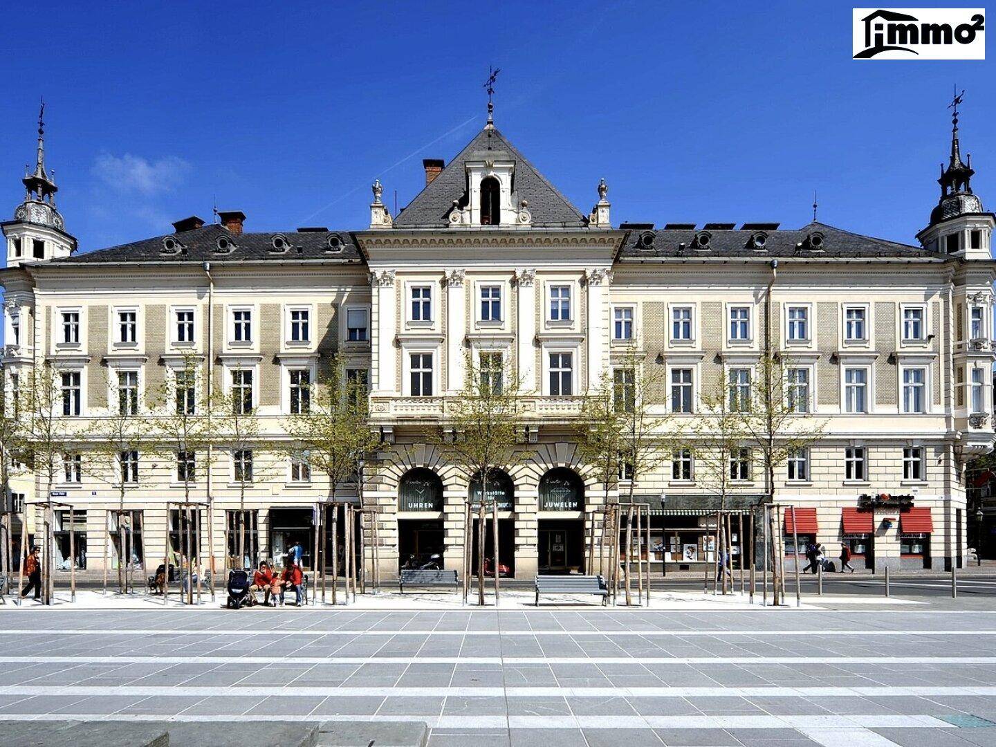 Historisches Gebäude mit klassischer Architektur, vielen Fenstern und Geschäften im Erdgeschoss an einem sonnigen Tag.