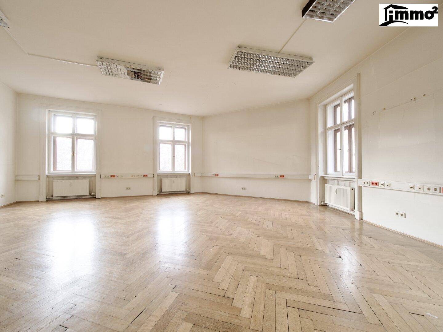 Großer, heller Büroraum mit Fischgrätparkett, drei hohen Fenstern und viel Tageslicht.