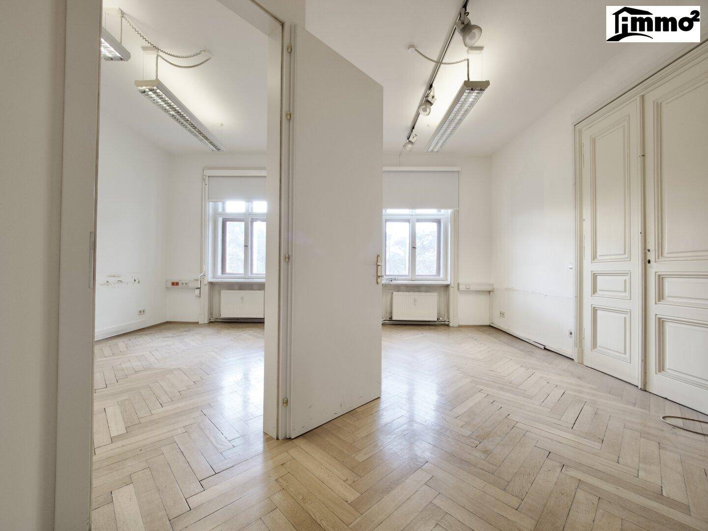 Heller Büroraum mit Fischgrätparkett, zwei Fenstern und einer offenen Tür zu einem weiteren Zimmer.